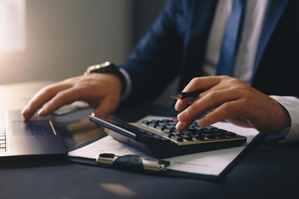Business man using a calculator at his desk, next to a laptop.